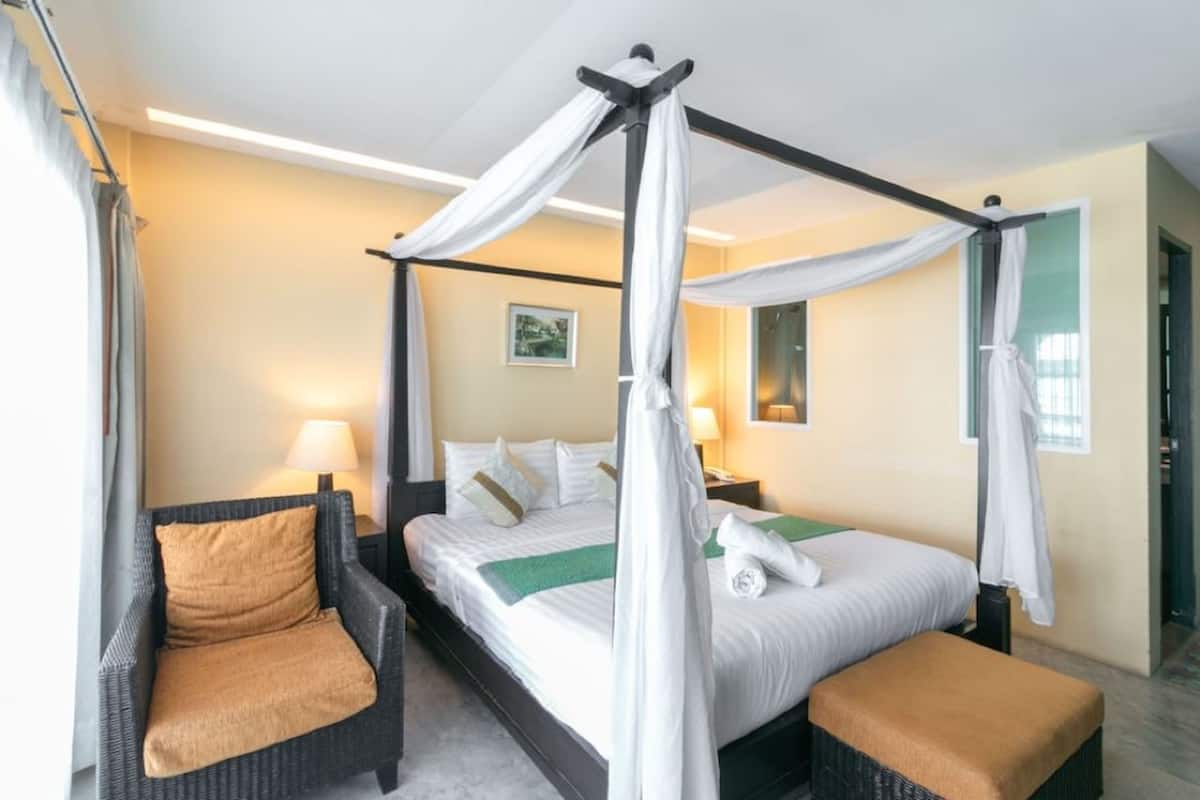 Una cama con dosel y cuatro columnas con ropa de cama blanca, un plaid verde y un cojín marrón en una silla de mimbre oscura.