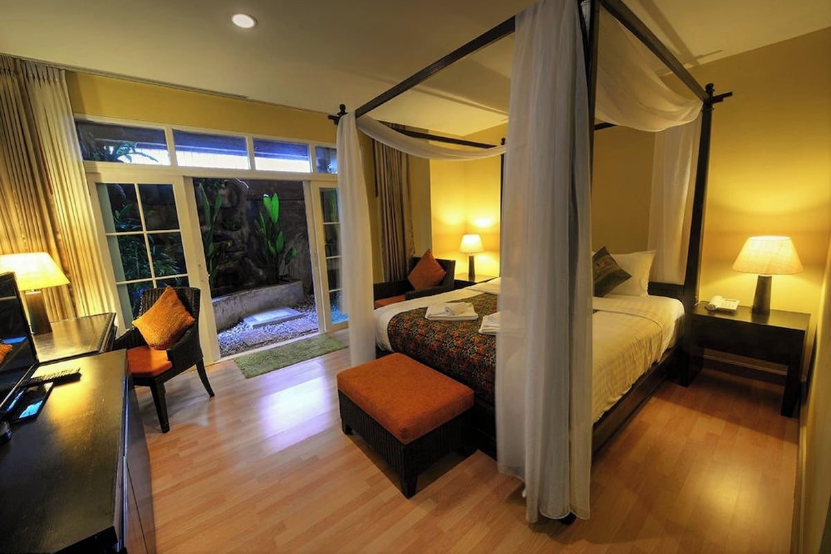 A four-poster bed with a canopy, a desk with a lamp, and a view of a tropical garden through large windows.
