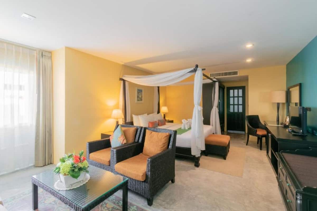 Habitación de hotel con cama con dosel, zona de estar con sofá y sillones, una mesita con un jarrón de flores y un televisor en una cómoda.