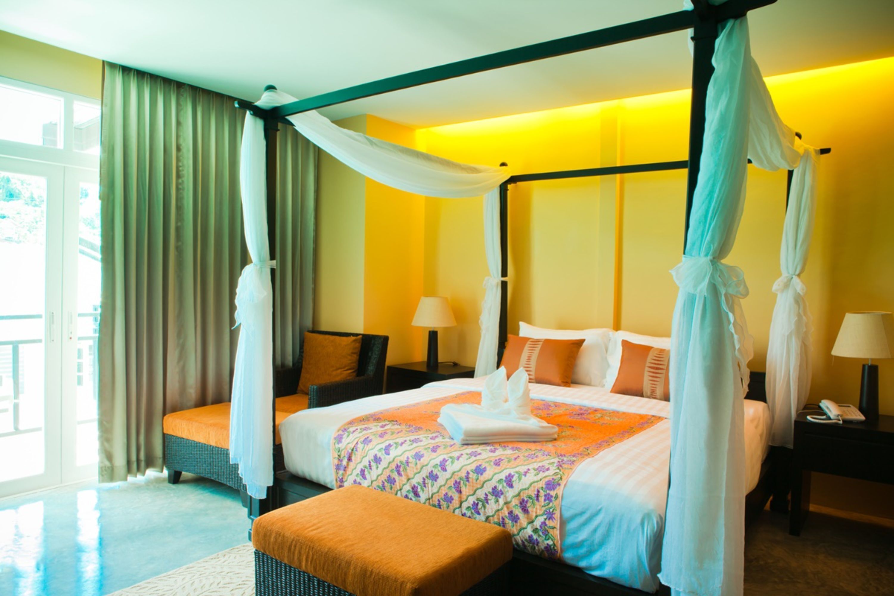 A four-poster canopy bed with a floral bedspread, a bedside table with a lamp, and a view of the outdoors through a large window.