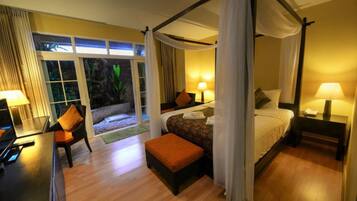 A four-poster bed with a canopy, a desk with a lamp, and a view of a tropical garden through large windows.