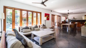 A spacious living area with a large sofa, a coffee table, and a dining table with chairs. There are ceiling fans and a kitchen area visible in the background. - Villa Martine (Seminyak)