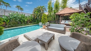 A poolside area with a wooden table, wicker furniture, and a covered seating area. - Villa Martine (Seminyak)