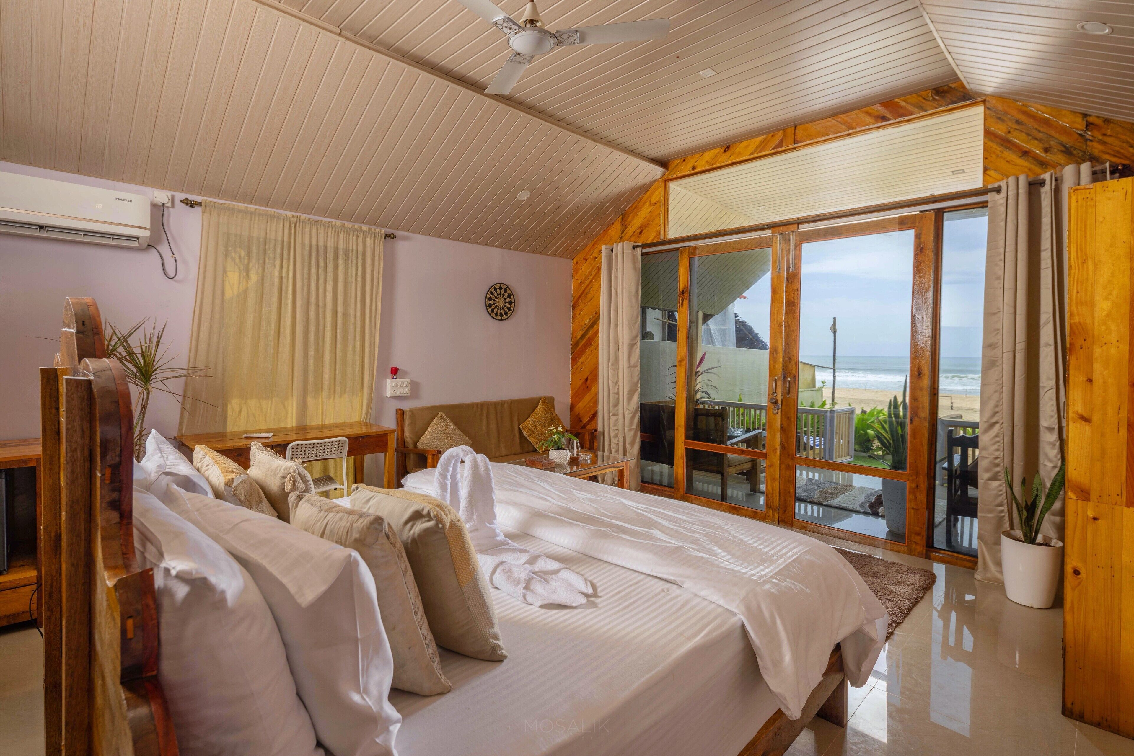 A bedroom with a large bed, a desk, a chair, and a view of the beach through a sliding door.
