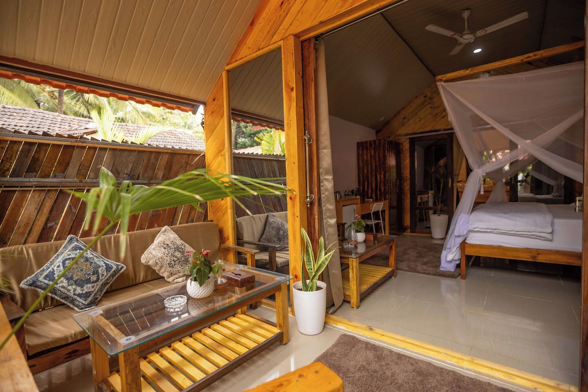 A wooden interior with a glass table, a sofa with cushions, a bed with a canopy, and a potted plant.