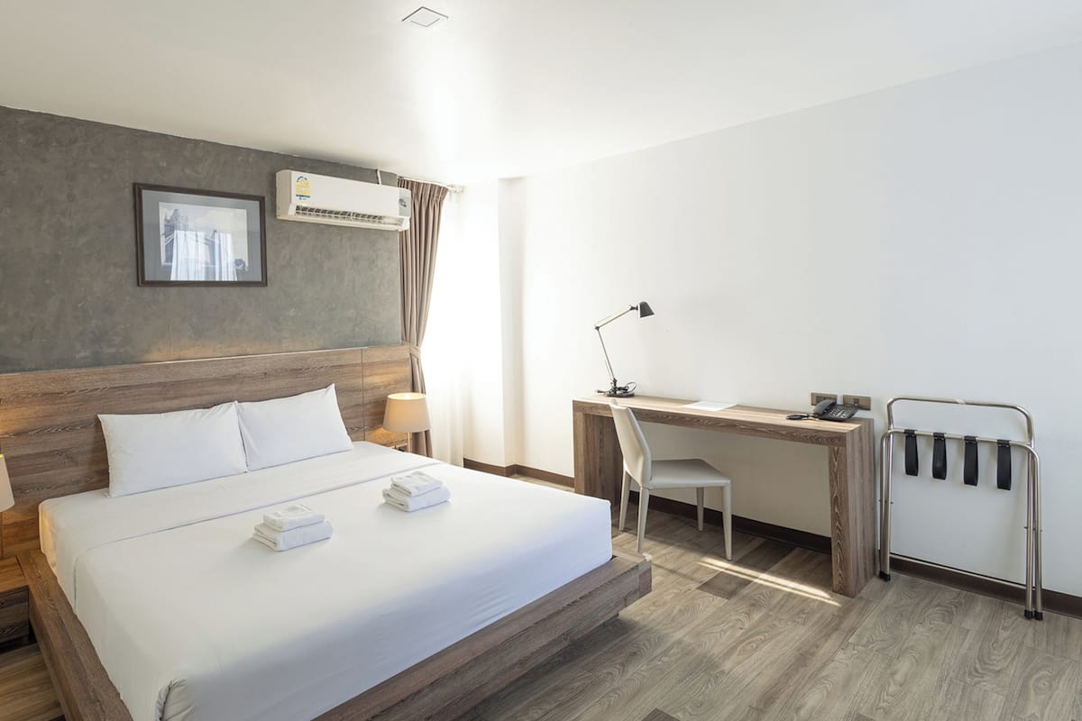 A hotel room with a bed, desk, chair, lamp, air conditioning unit, and a framed picture on the wall.