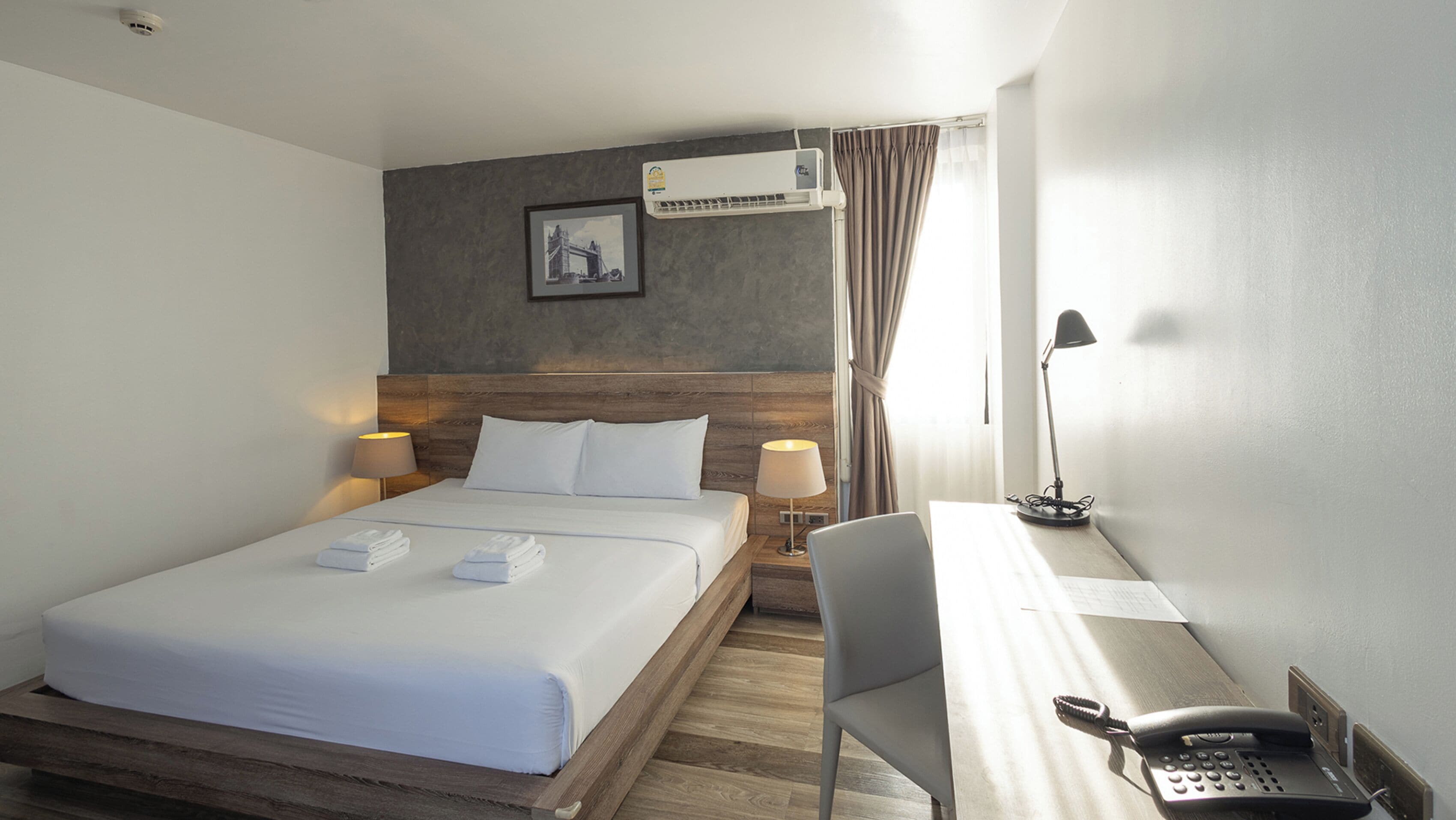 A hotel room with a bed, two bedside lamps, a desk with a telephone, and an air conditioning unit.