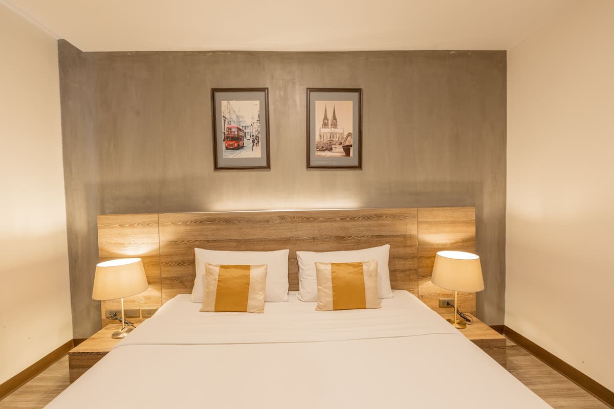 A hotel room with a wooden headboard, two framed pictures on the wall, a bed with white linens and gold pillows, and two bedside lamps.