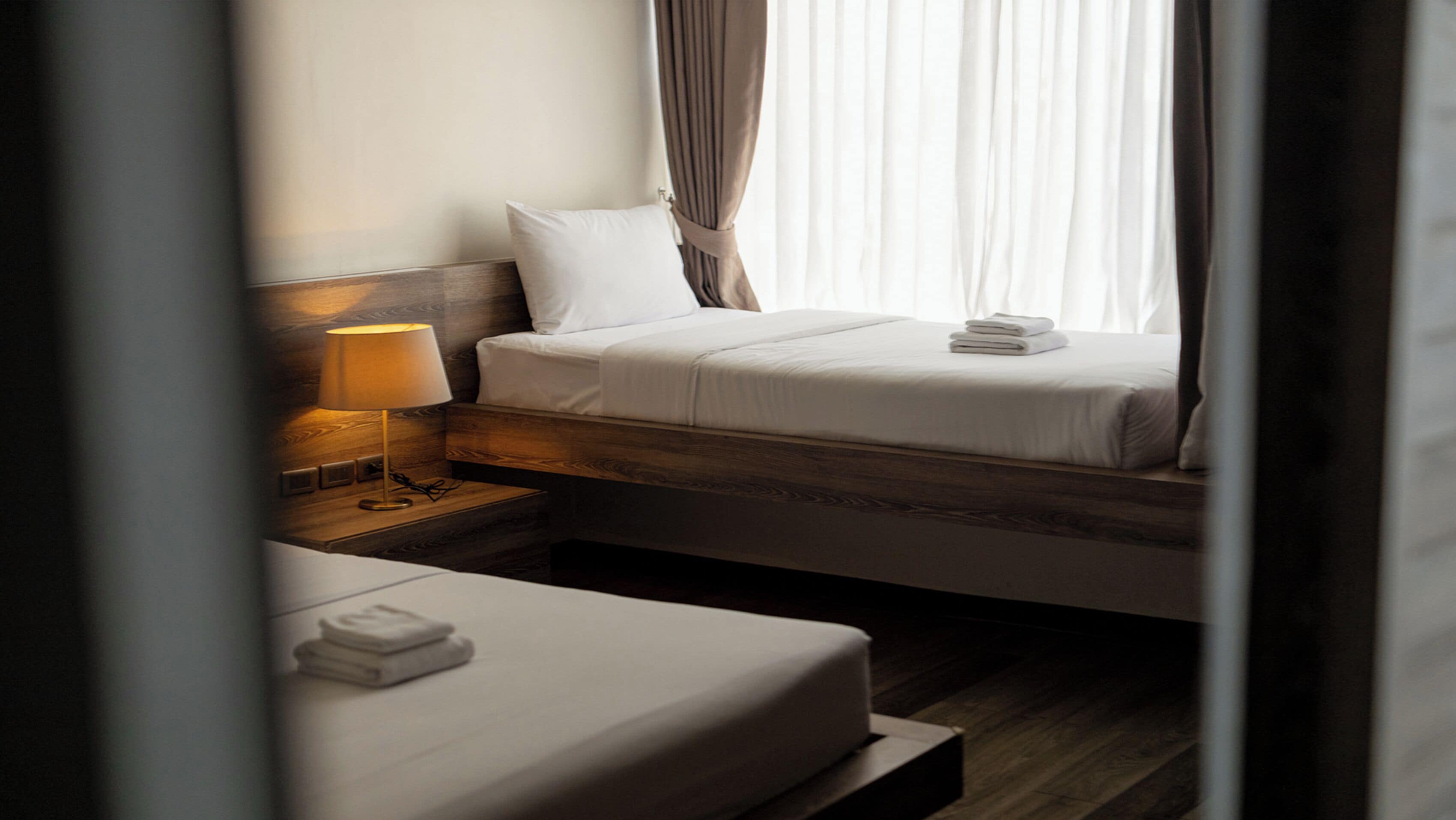 Ein Hotelzimmer mit einem Bett, einem Nachttisch mit Lampe und Blick ins Freie durch Vorhänge.