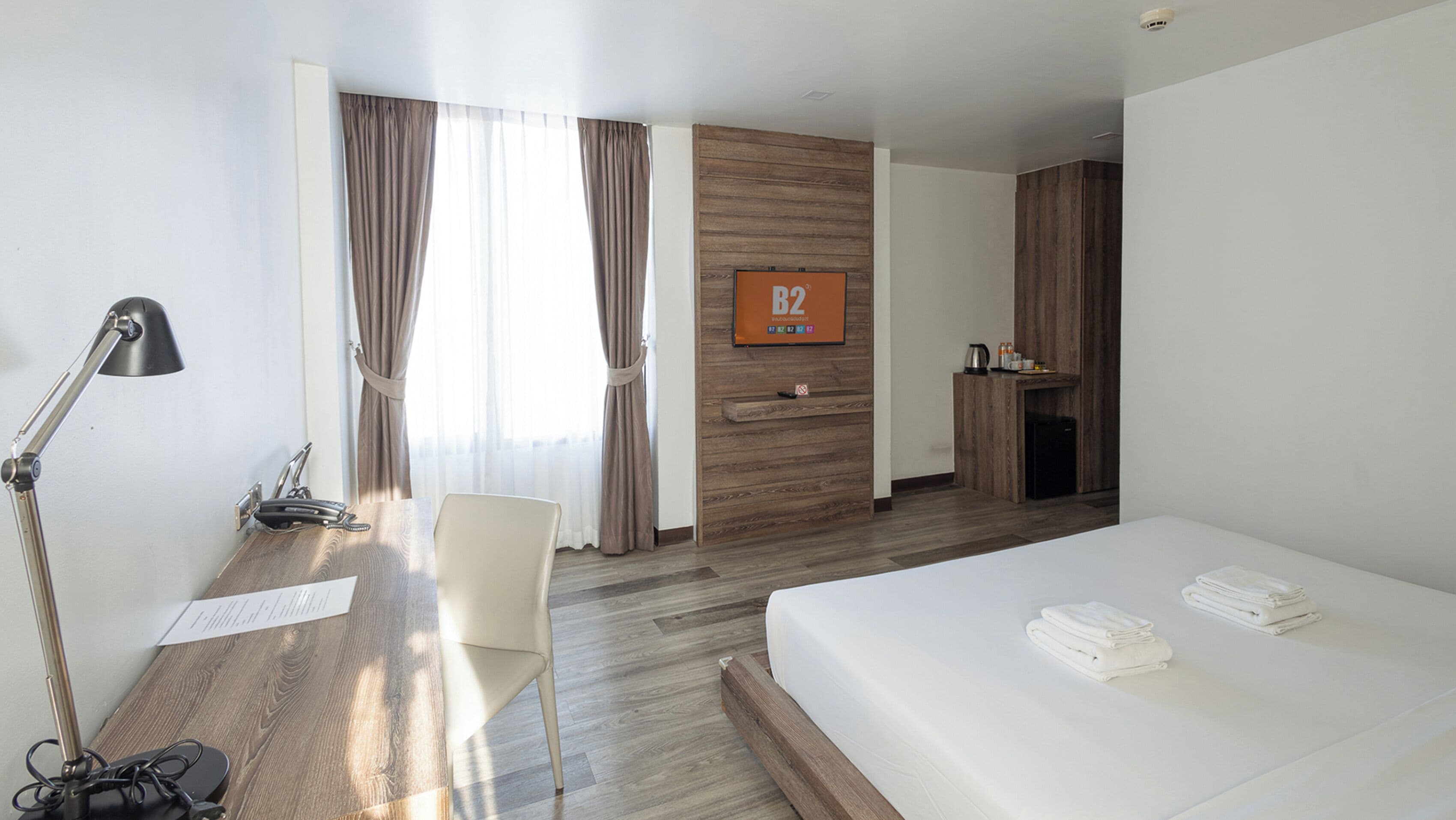 Ein Hotelzimmer mit einem Bett, einem Schreibtisch mit Lampe, einem Sessel, einer Holzverkleidung, einer kleinen Küchenzeile und einem Fenster mit Vorhängen.