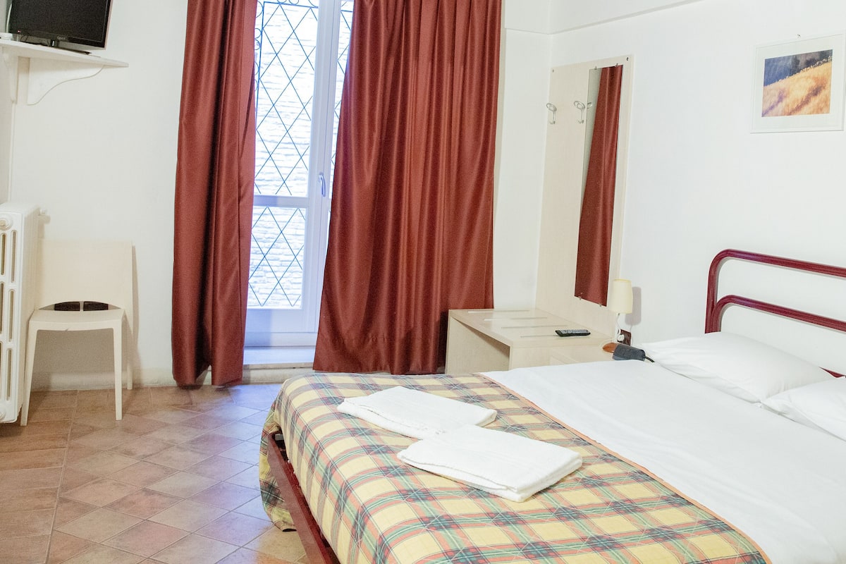Habitación de hotel con cama, escritorio, silla, radiador y televisor montado en la pared.