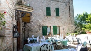 An outdoor dining area with tables and chairs, a brick building with green shutters, and a rustic ambiance. - Palazzo Meraviglia - Albergo Diffuso (Corinaldo)