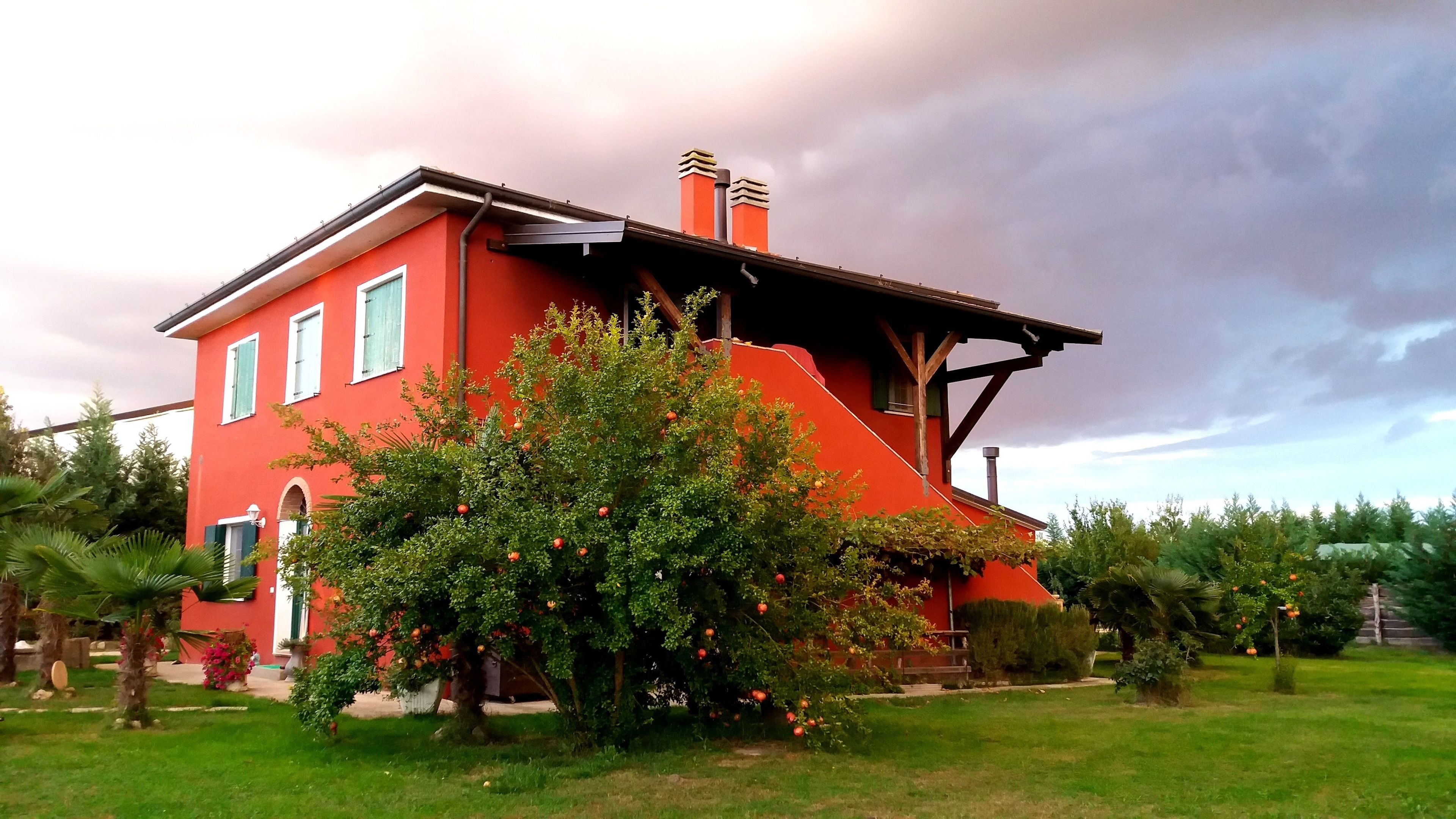 A red house with a sloped roof and two chimneys, surrounded by a well-maintained garden with various plants and a tree bearing fruit.