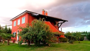 A red house with a sloped roof and two chimneys, surrounded by a well-maintained garden with various plants and a tree bearing fruit.