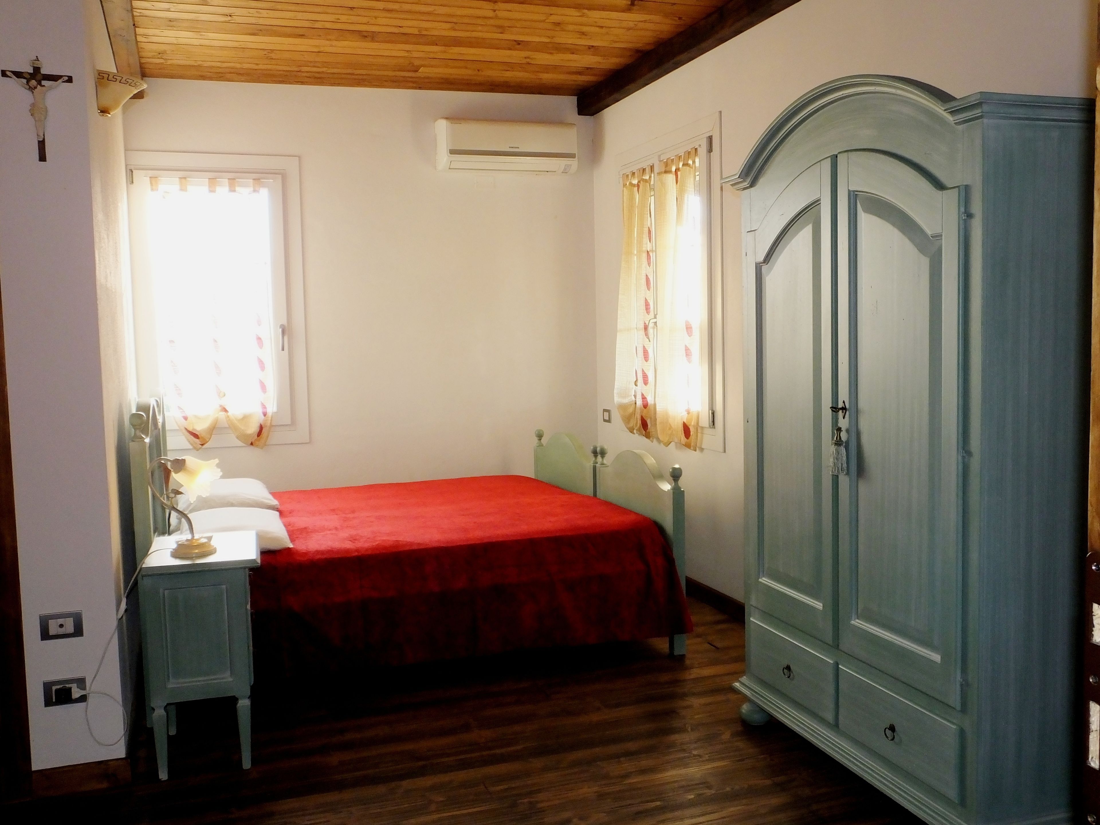 A bedroom with a wooden ceiling, a bed with a red bedspread, a nightstand, a wardrobe, and a window with curtains.