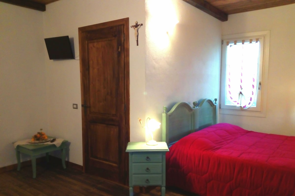A bedroom with a wooden door, a bedside table with a lamp, a bed with a red blanket, and a window with curtains.