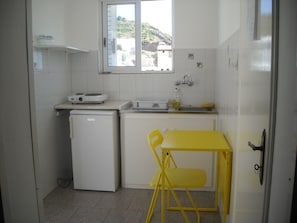 A compact kitchen with a small refrigerator, a sink, and a window. - Apostolakis Rooms (Ikaria)