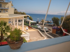 A coastal view with a building, a green bus, and a white car parked near the water. - Apostolakis Rooms (Ikaria)