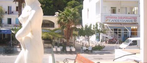 A public square with a white statue, a wooden bench, and a building with a sign in Greek.