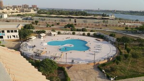 A swimming pool with a unique shape, surrounded by a paved area with outdoor seating and a view of a river. - Minya Hotels of the Armed Forces (El Minya)