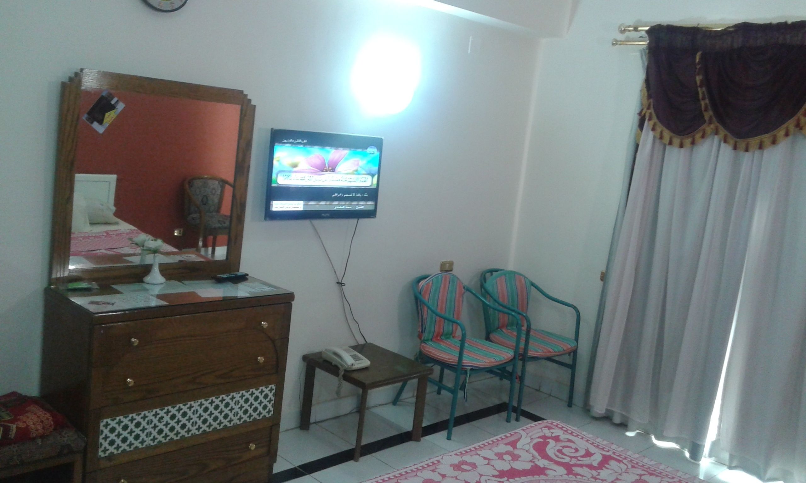 A room with a wooden dresser, a small table with a phone, two striped chairs, a television on the wall, and a patterned rug.
