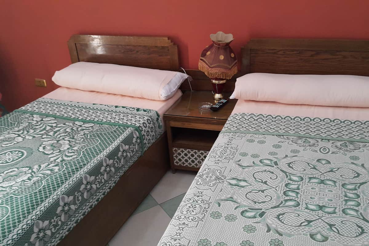 A room with two single beds with floral-patterned bedding, a wooden nightstand with a vase and remote control, and a red wall.