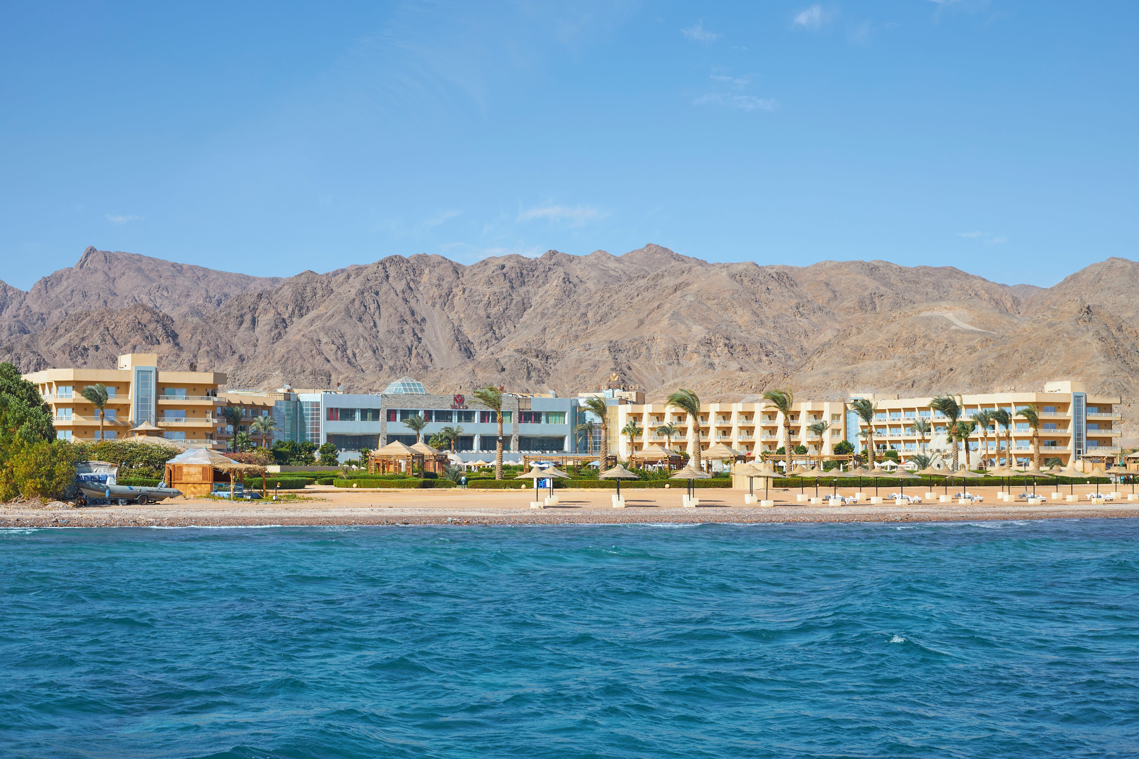 Un hotel frente al mar con varias edificaciones, una playa y montañas al fondo.
