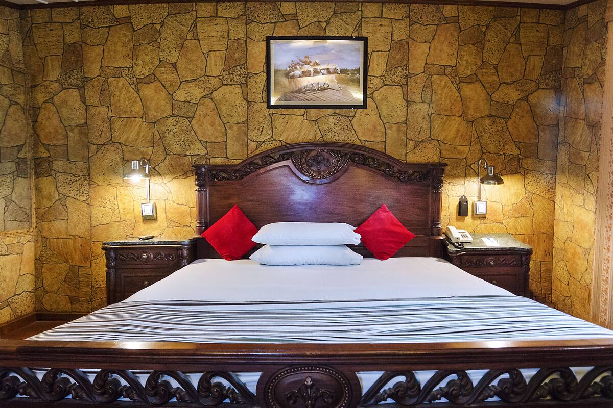 A hotel room with a stone-patterned wall, a large bed with white and red pillows, and two bedside tables with lamps.