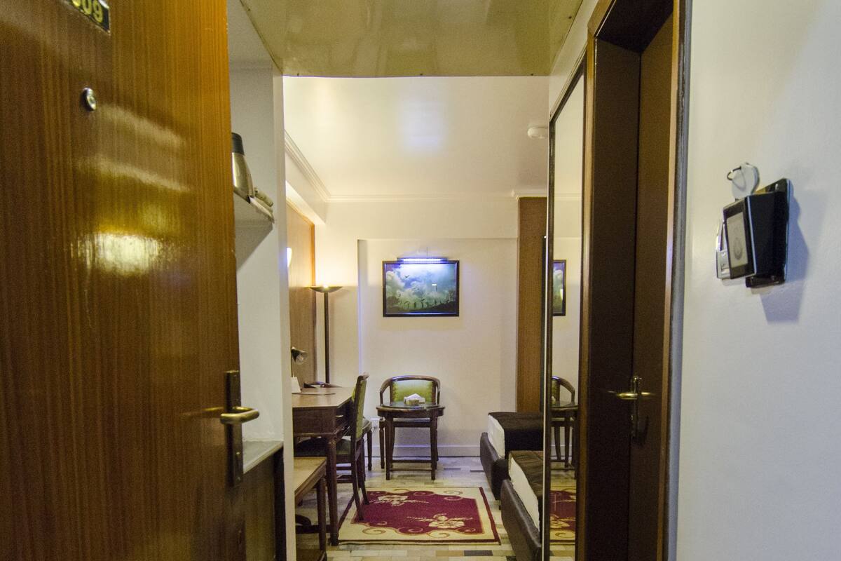 A hallway with a wooden door on the left, a dining area with a table and chairs in the center, and a bed with a red rug in the background.