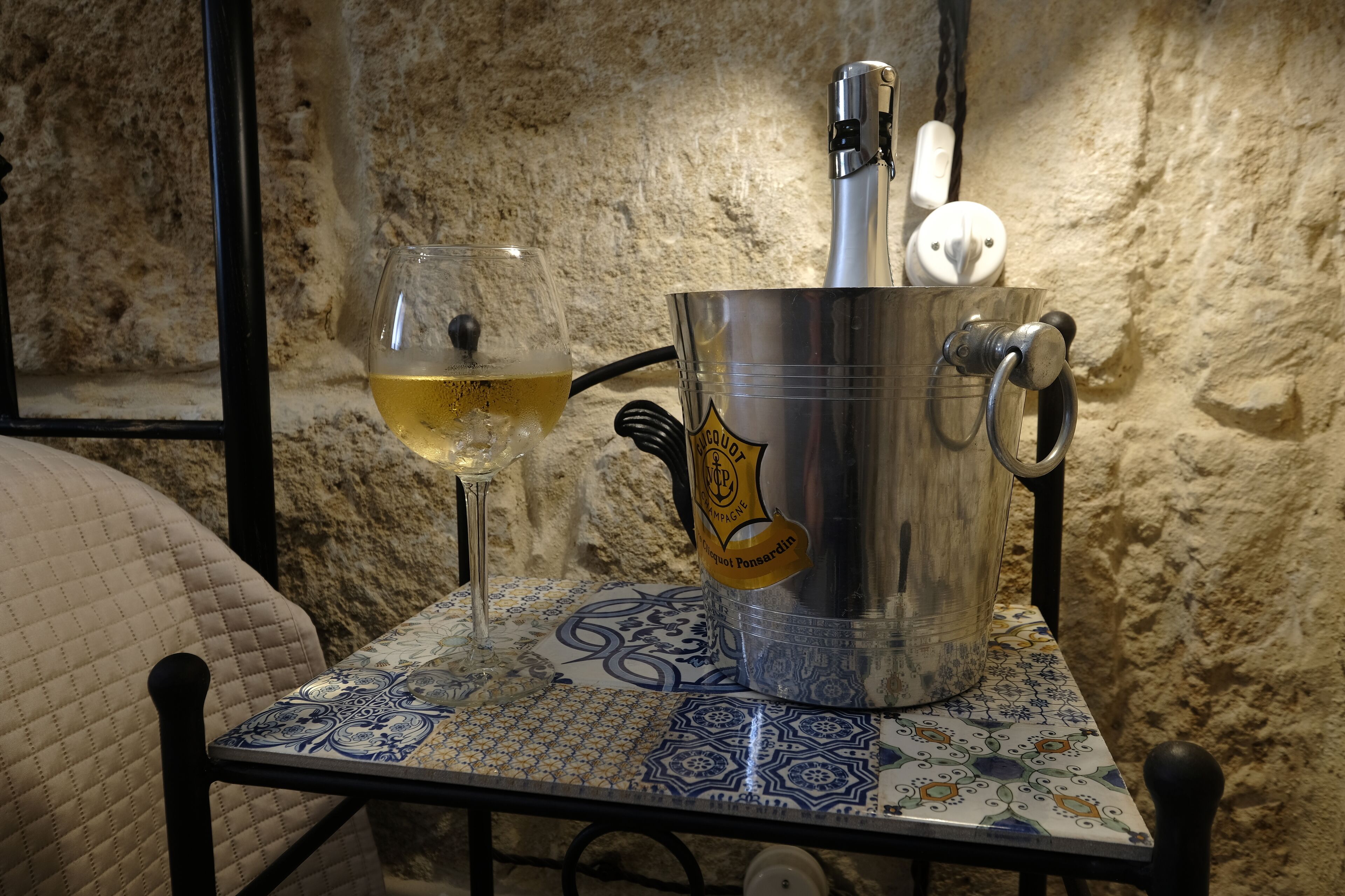 Une table avec un verre de vin blanc et un récipient en métal portant une étiquette, le tout placé devant un mur en pierre à la texture marquée.
