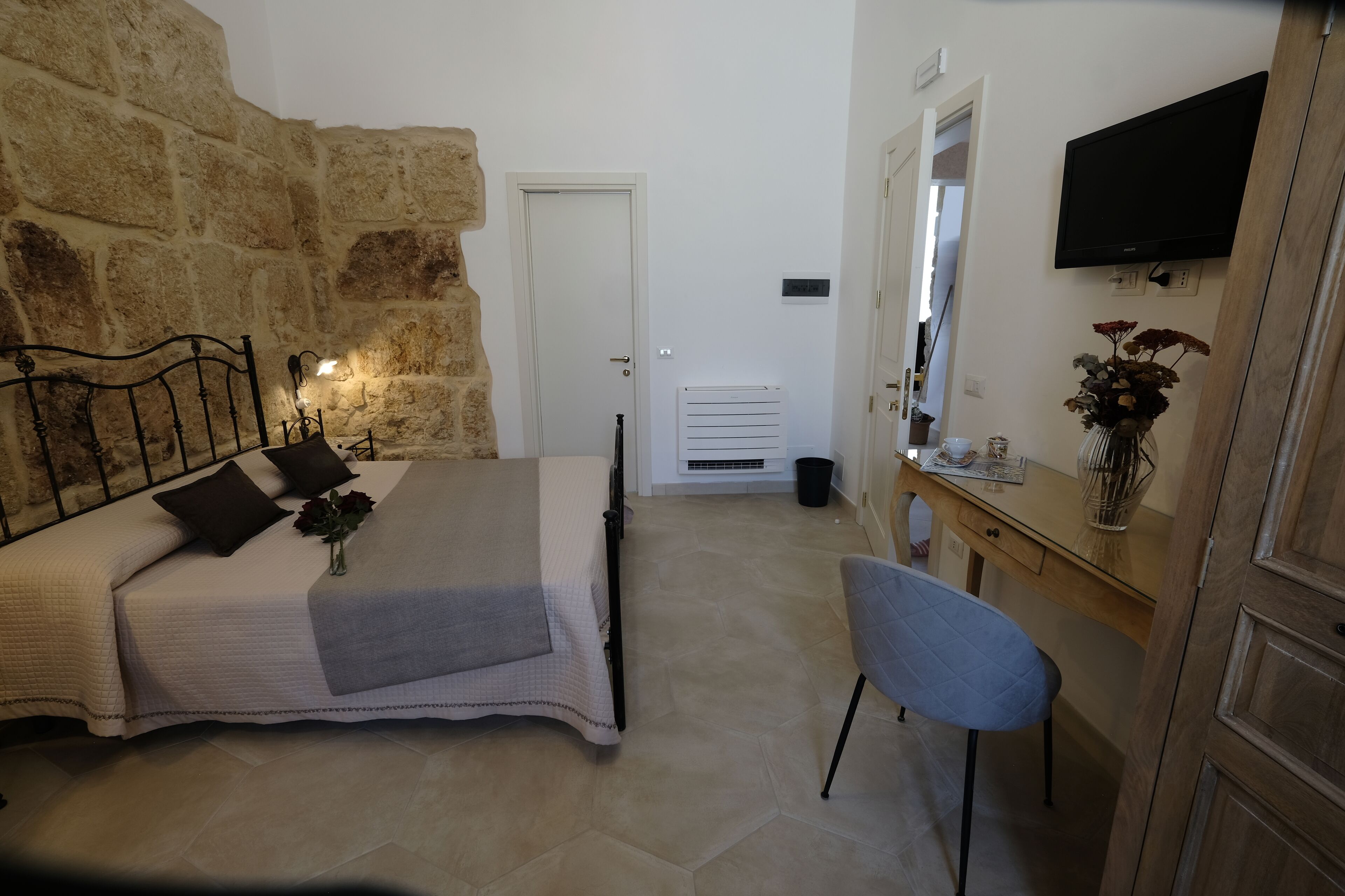 Un dormitorio con pared de piedra, una cama con cabecera, una mesita de noche, una silla, un escritorio, un espejo y un televisor montado en la pared.