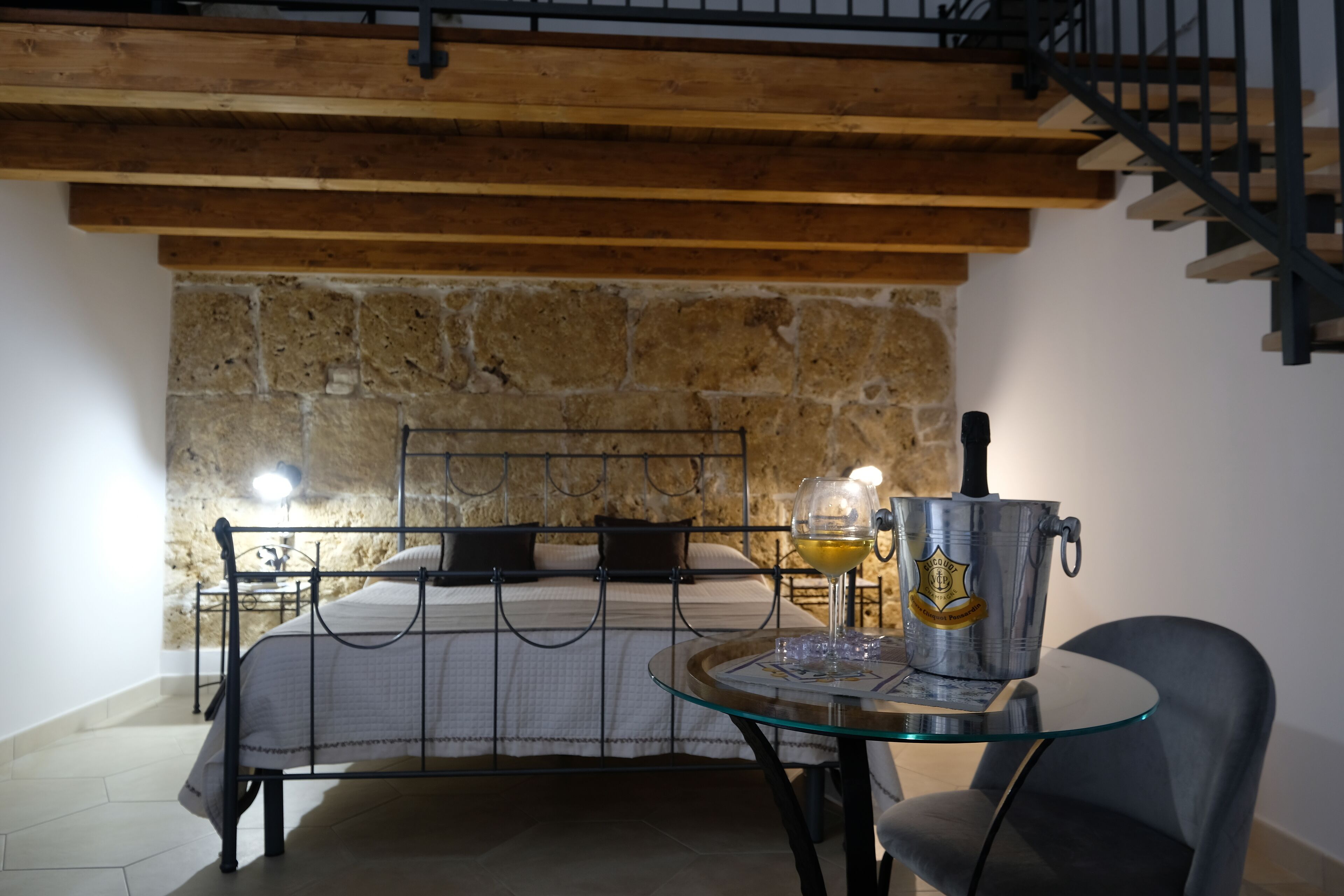 A bedroom with a stone wall, a bed with black metal headboard, a glass table with a wine glass and a metal bucket, and a staircase with a metal railing.