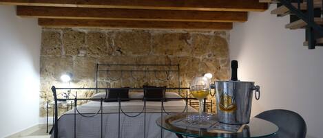 A bedroom with a stone wall, a bed with black metal headboard, a glass table with a wine glass and a metal bucket, and a staircase with a metal railing.
