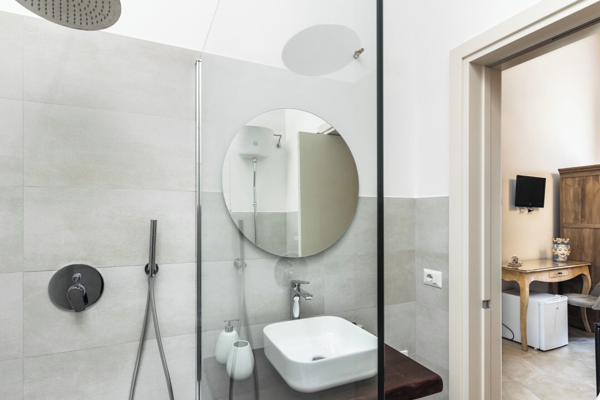 A modern bathroom with a glass shower enclosure, a round mirror, a white sink, and a wooden vanity with a television.