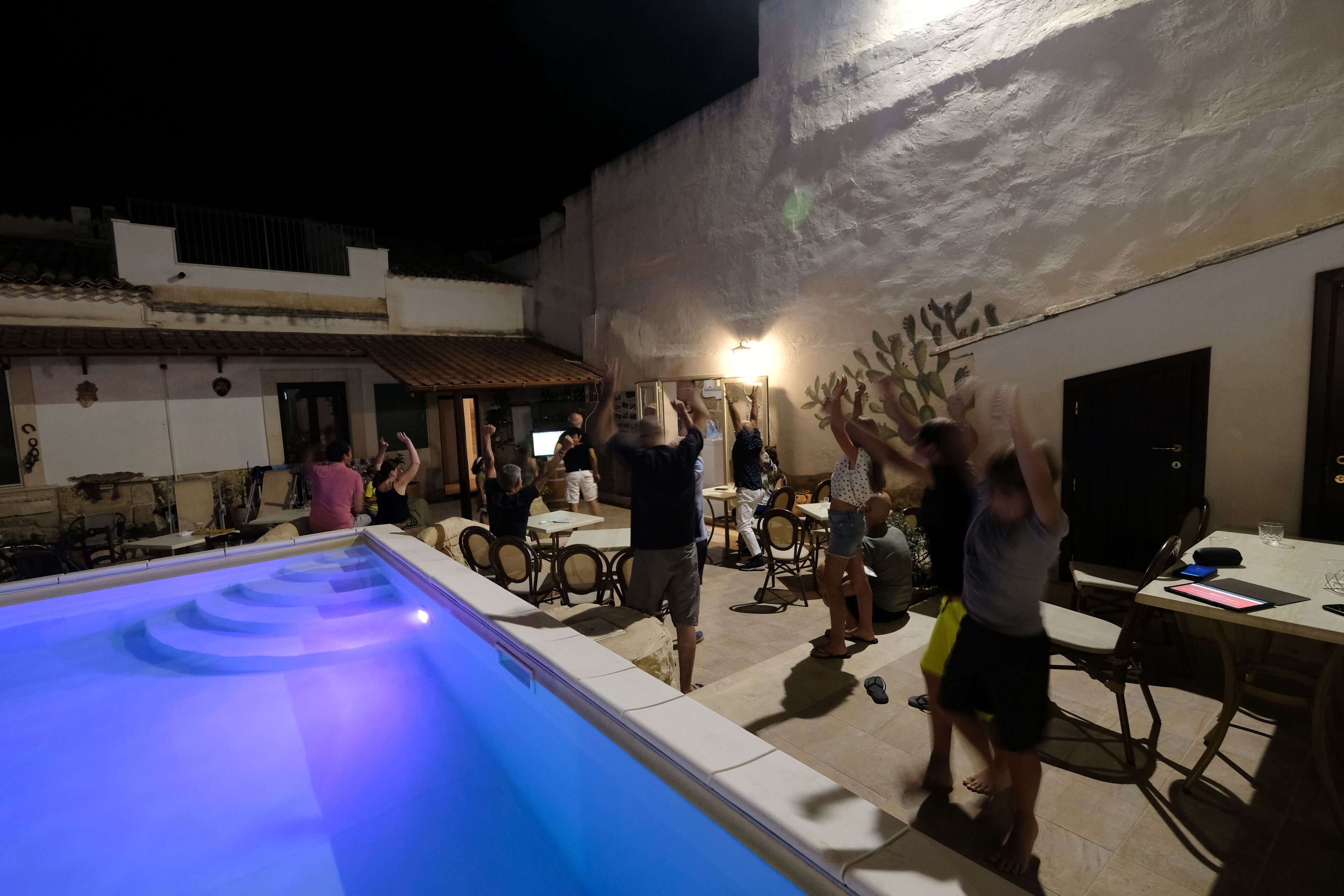 Un área de piscina al aire libre con una fuente iluminada en azul, rodeada de mesas y sillas, y una pared texturizada al fondo.