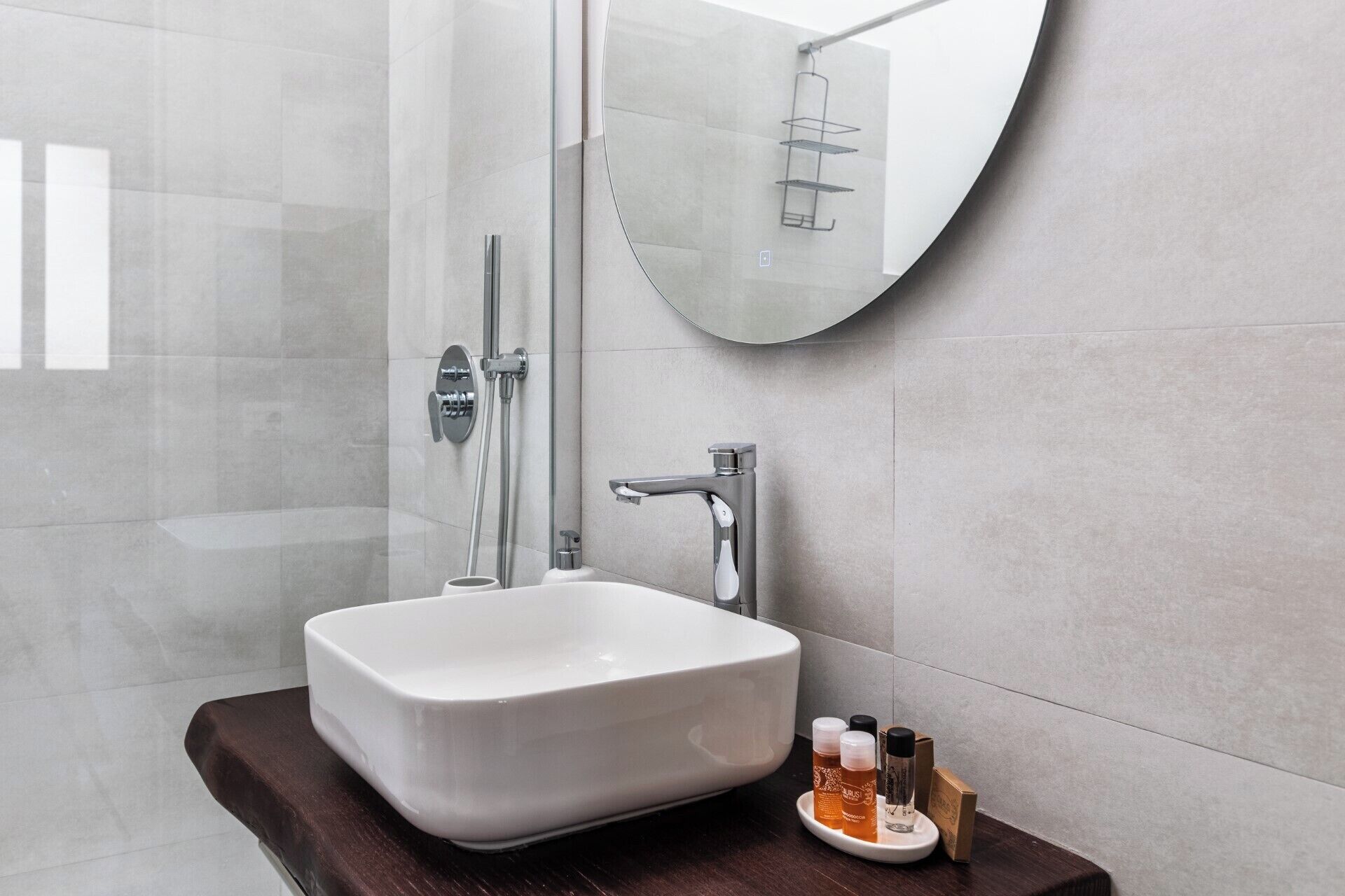 Un baño moderno con ducha envuelta en vidrio, lavamanos blanco y espejo redondo.