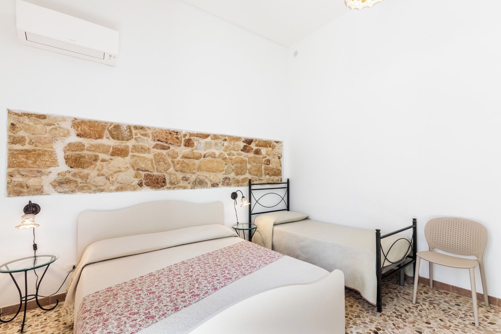 A bedroom with a stone wall, two beds, a chair, and a small table.