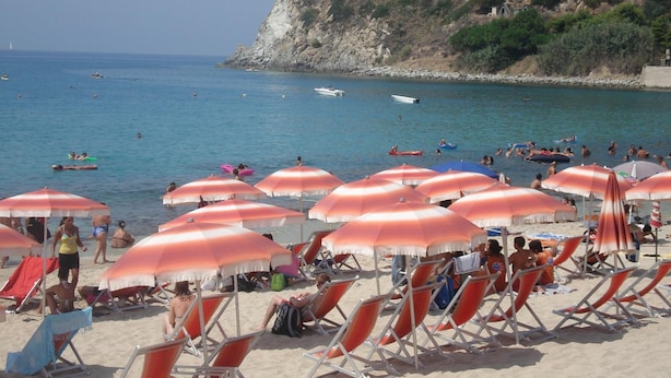 A beach with rows of sun loungers and umbrellas, a rocky hillside in the background, and boats on the water.