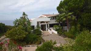 A white building with a red-tiled roof, surrounded by lush greenery and a stone pathway leading to the entrance.