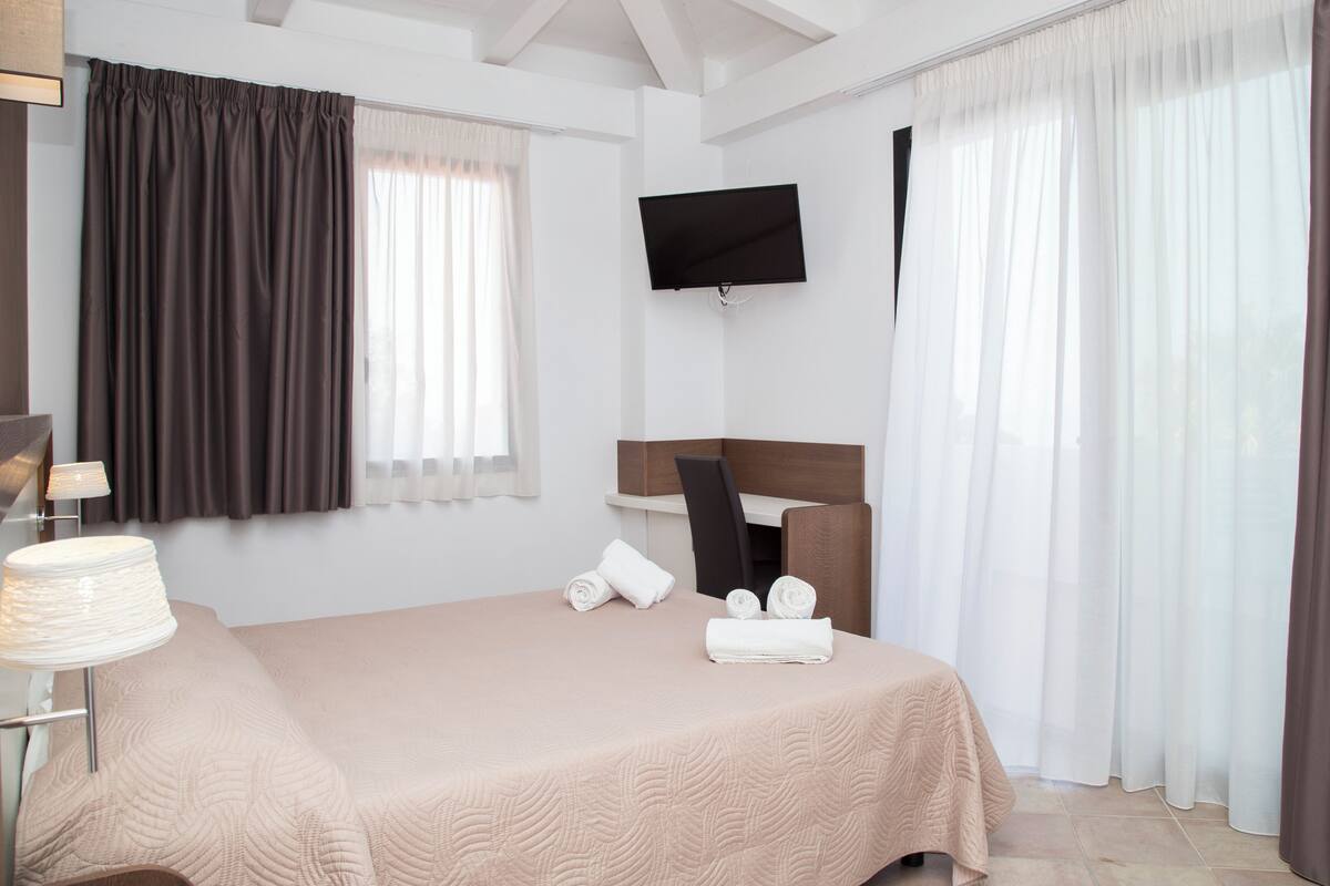 Una habitación de hotel con cama, televisor montado en la pared y ventana con cortinas.