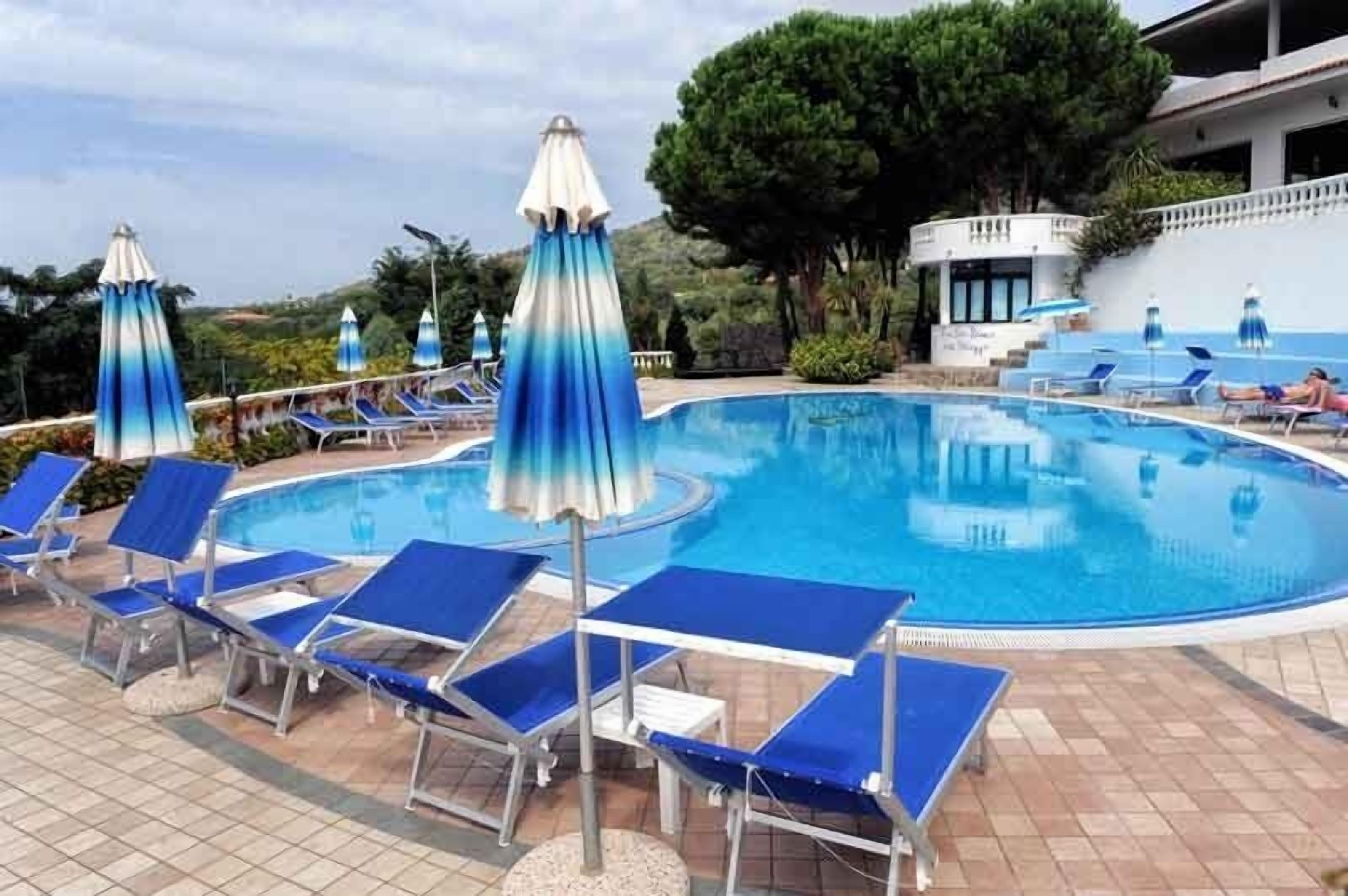 A swimming pool with blue lounge chairs and umbrellas, surrounded by trees and a building.