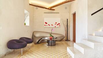 Un intérieur moderne avec un canapé incurvé, deux fauteuils violets, une table basse ornée d’un vase, un escalier et un sol à motifs.