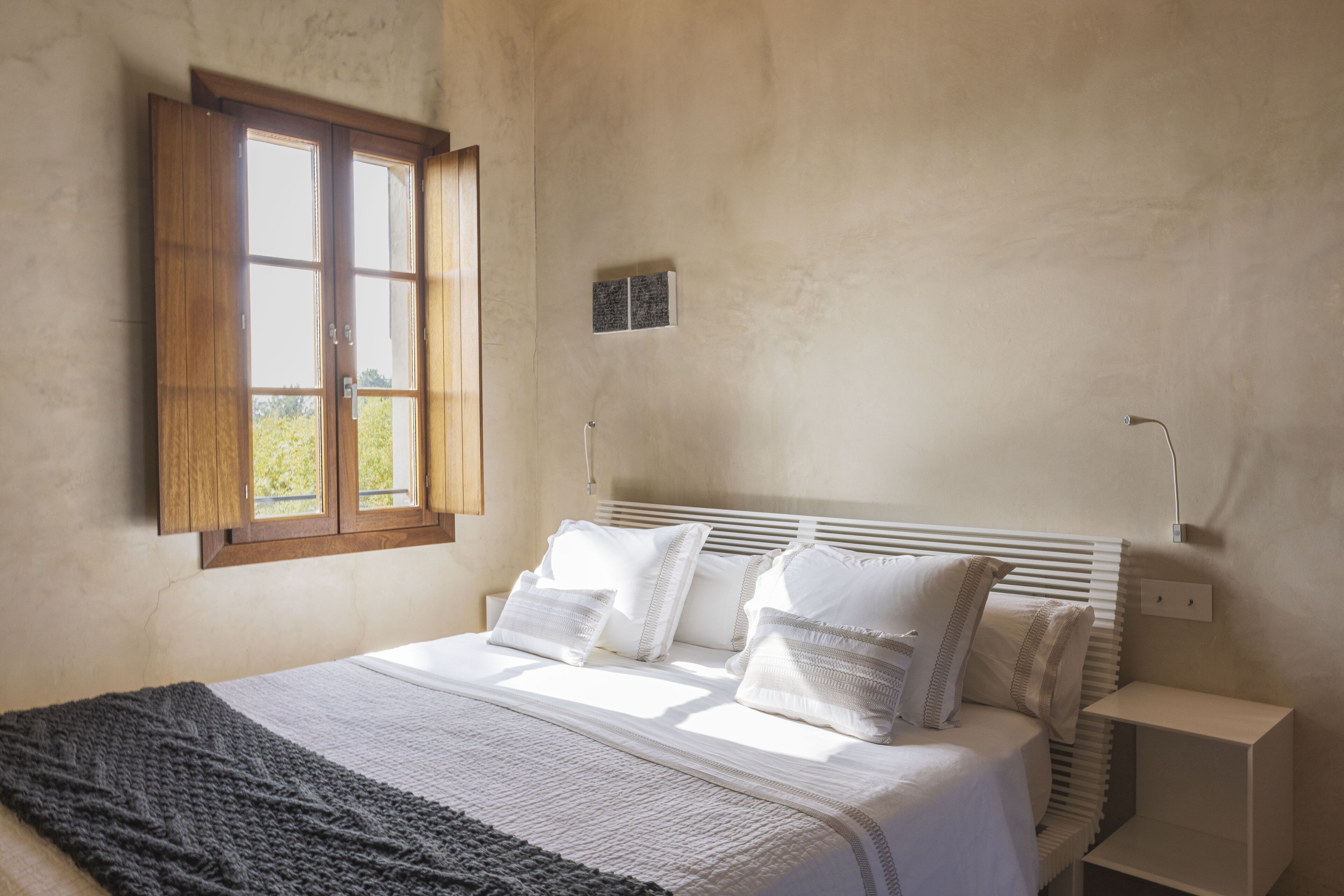 Un dormitorio con cama, ventana, mesita de noche y lámpara de pared.