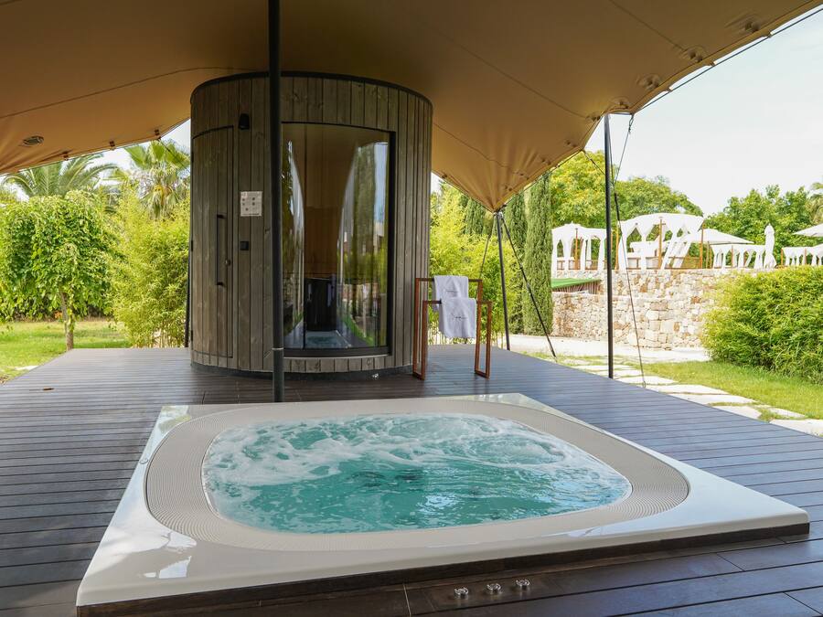 Ein Whirlpool unter einer Markise, umgeben von einer Holzterrasse, mit einer Steinmauer und Grünpflanzen im Hintergrund.