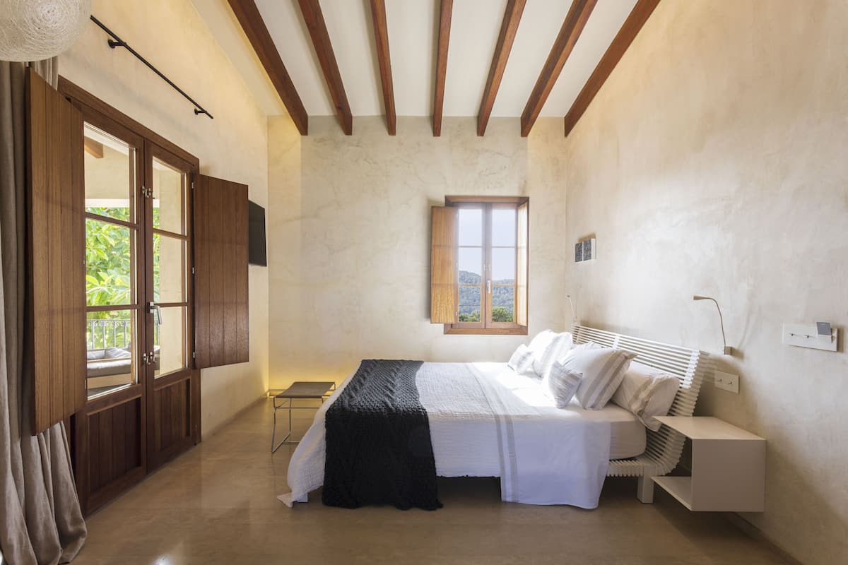 A bedroom with a bed, bedside table, lamp, window, and wooden ceiling beams.