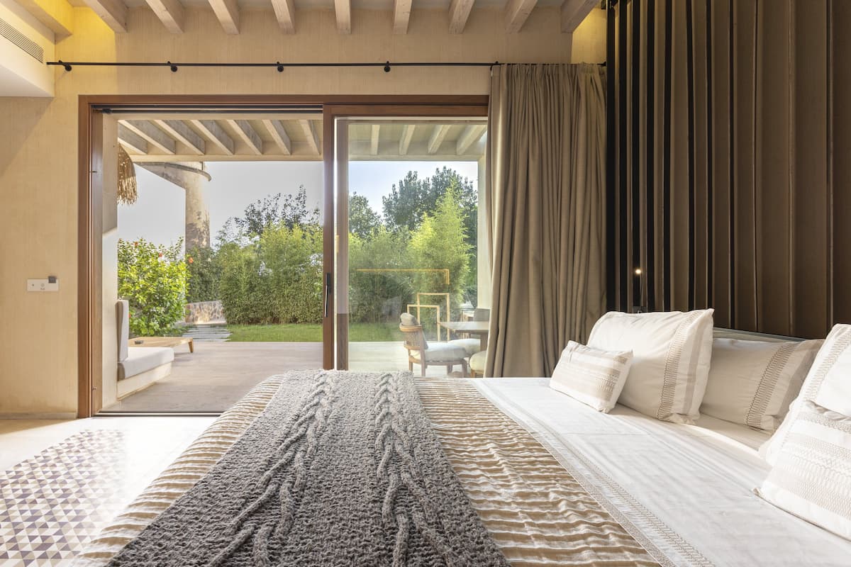 A bedroom with a large bed, a view of a garden, and a sliding glass door leading to an outdoor seating area.