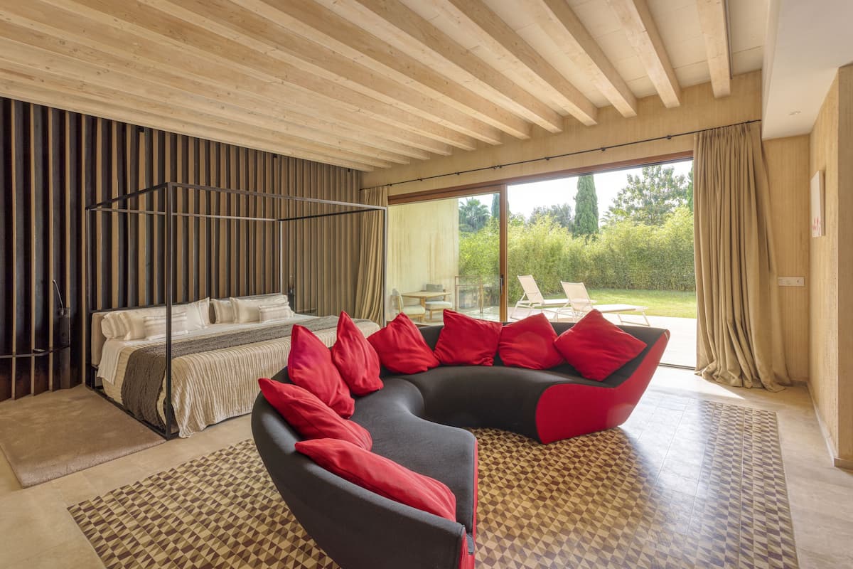 A modern bedroom with a curved black sofa, a bed with red pillows, and a large window offering a view of the outdoors.