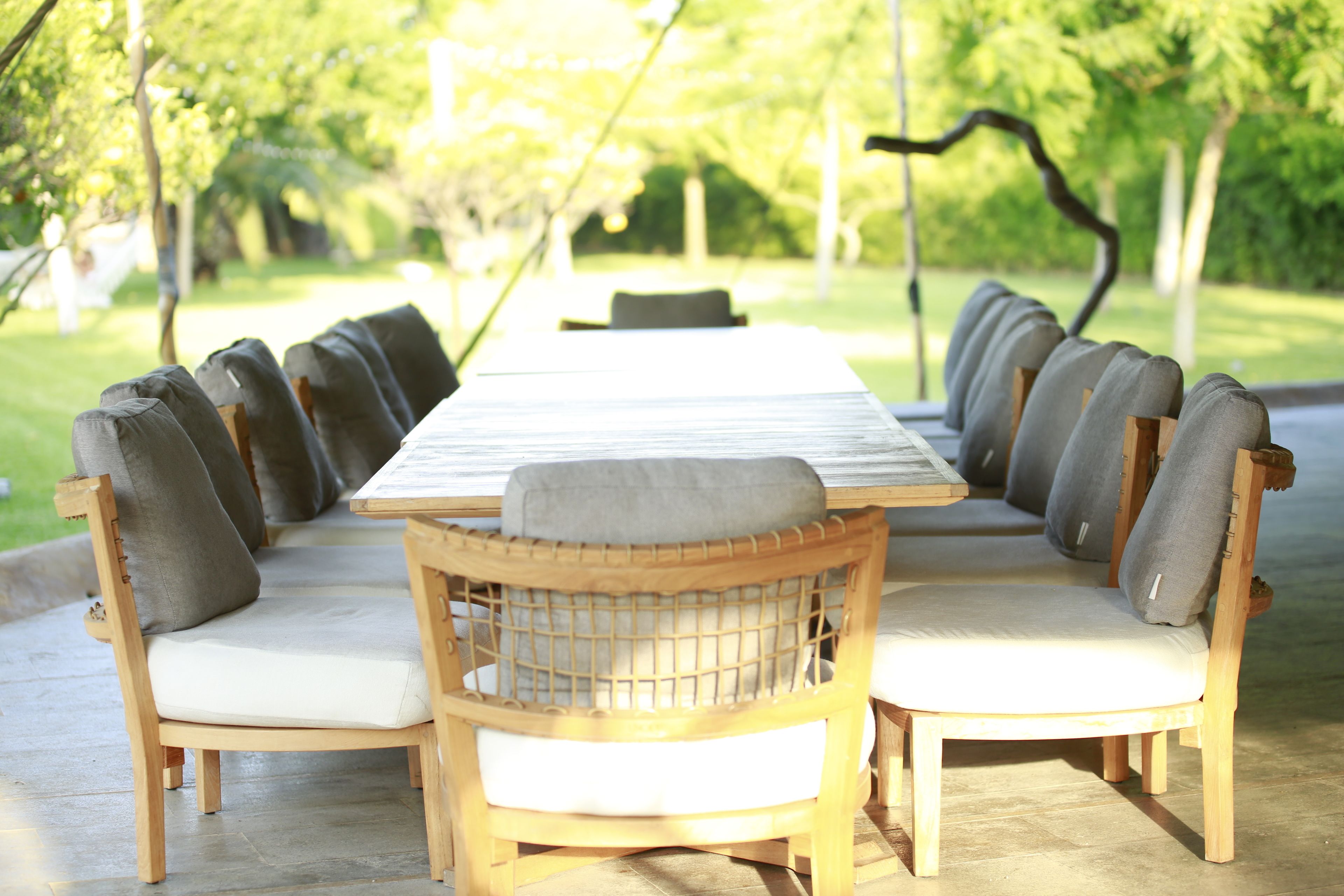 A long wooden table with cushioned chairs, set outdoors with a view of trees and a building in the background.