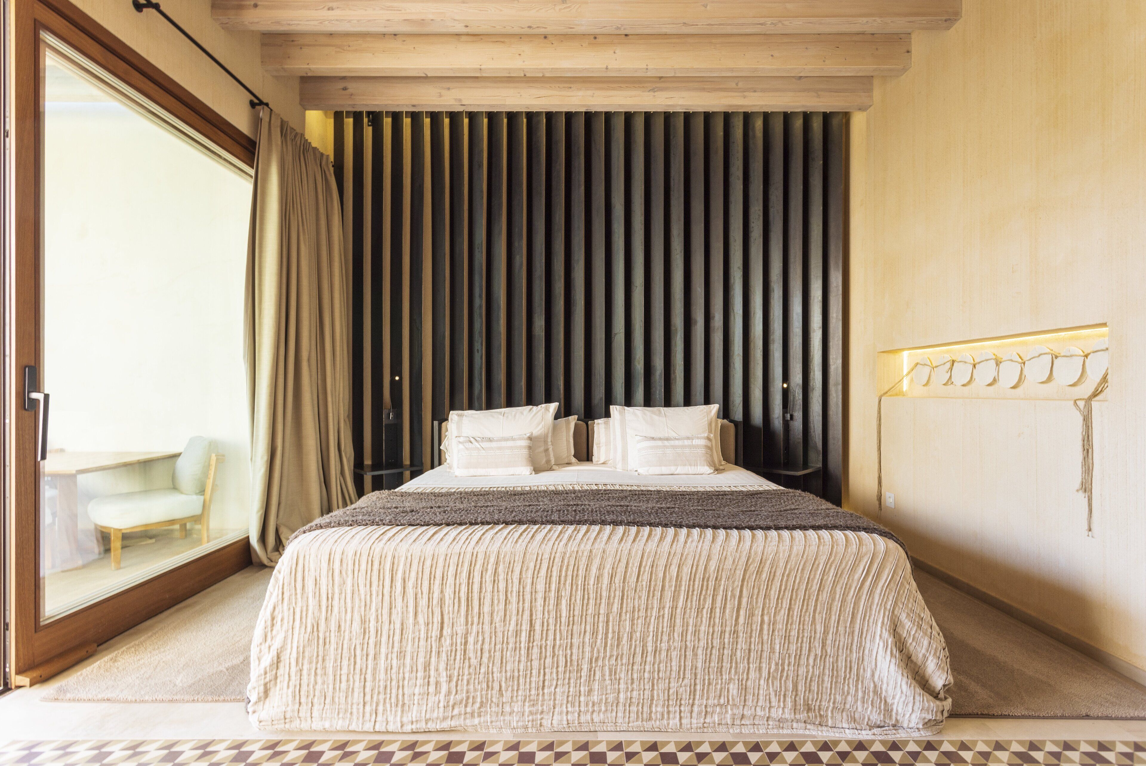 Un dormitorio con una cama grande, vistas al exterior a través de una puerta corredera, una pequeña silla y un diseño de pared único.