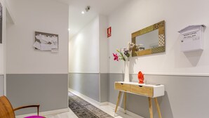 A hallway with a wooden chair, a white console table with a vase of flowers, a mirror with a geometric frame, and a key mail box. - Hostal Met Madrid (Madrid)